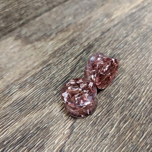 May include: Two round, clear, pink resin ear gauges with embedded copper flakes. The gauges are on a brown wooden surface.