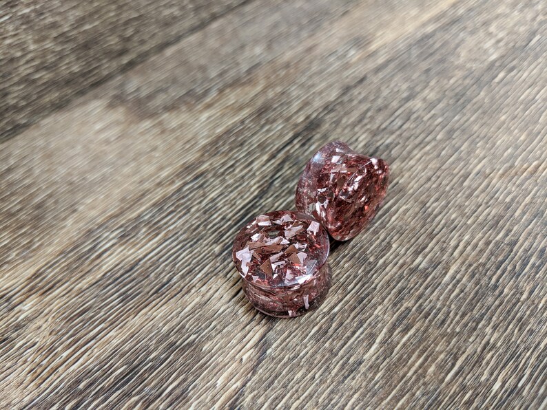 May include: Two round, clear, pink resin ear gauges with embedded copper flakes. The gauges are on a brown wooden surface.