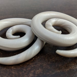 May include: A pair of white spiral ear gauges with a pearlescent finish. The gauges are made of a smooth, shiny material and have a swirling design.