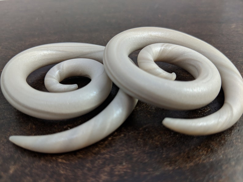 May include: A pair of white spiral ear gauges with a pearlescent finish. The gauges are made of a smooth, shiny material and have a swirling design.