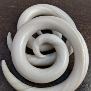 May include: A pair of white spiral ear gauges made of acrylic. The gauges have a pearlescent finish and are designed to stretch the earlobe.