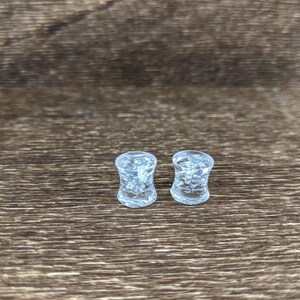 May include: Two clear glass ear gauges with a textured, bubbly surface. The gauges are on a brown wooden surface.