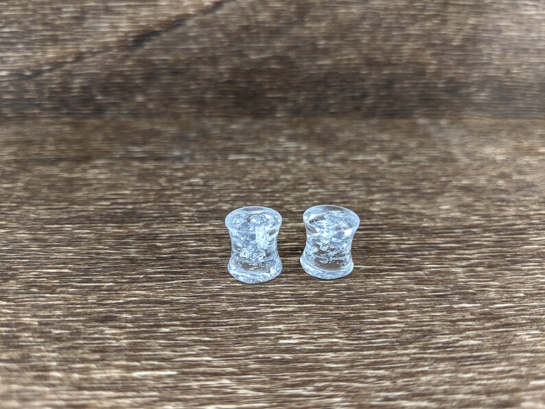 May include: Two clear glass ear gauges with a textured, bubbly surface. The gauges are on a brown wooden surface.