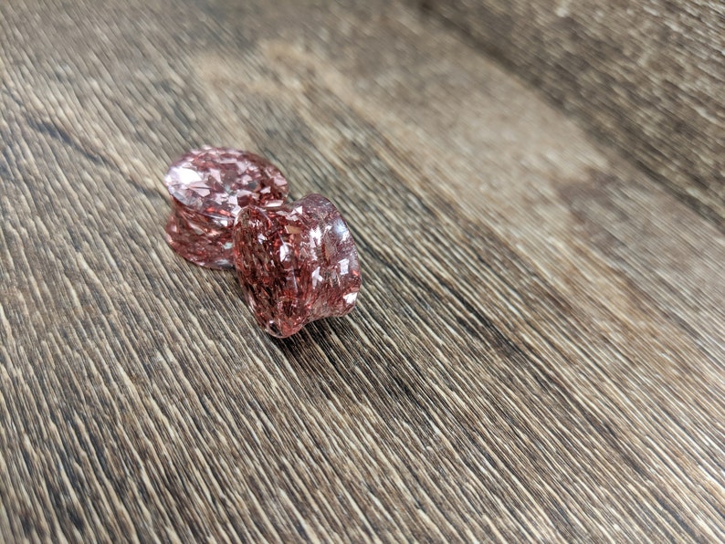 May include: Two pink and clear glass ear gauges with a glittery finish. The gauges are on a wood surface.
