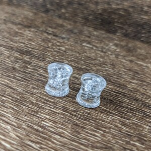 May include: Two clear glass ear gauges with a speckled, cloudy appearance. The gauges are on a brown wooden surface.