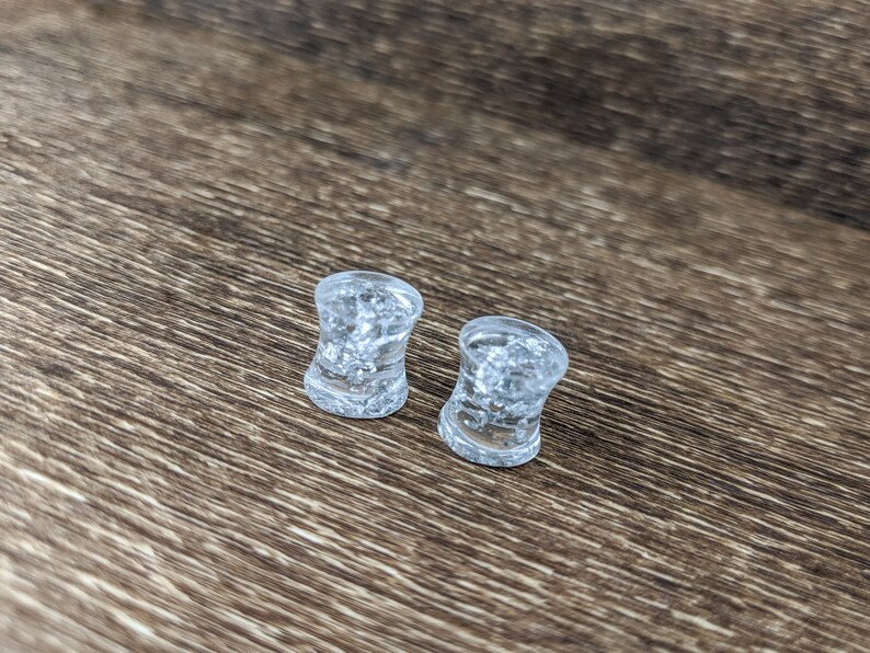 May include: Two clear glass ear gauges with a speckled, cloudy appearance. The gauges are on a brown wooden surface.