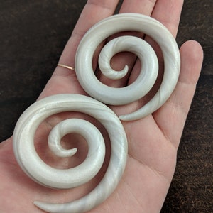 May include: A pair of white spiral ear gauges with a shimmery finish. The gauges are made of a smooth, polished material and have a subtle swirl pattern.