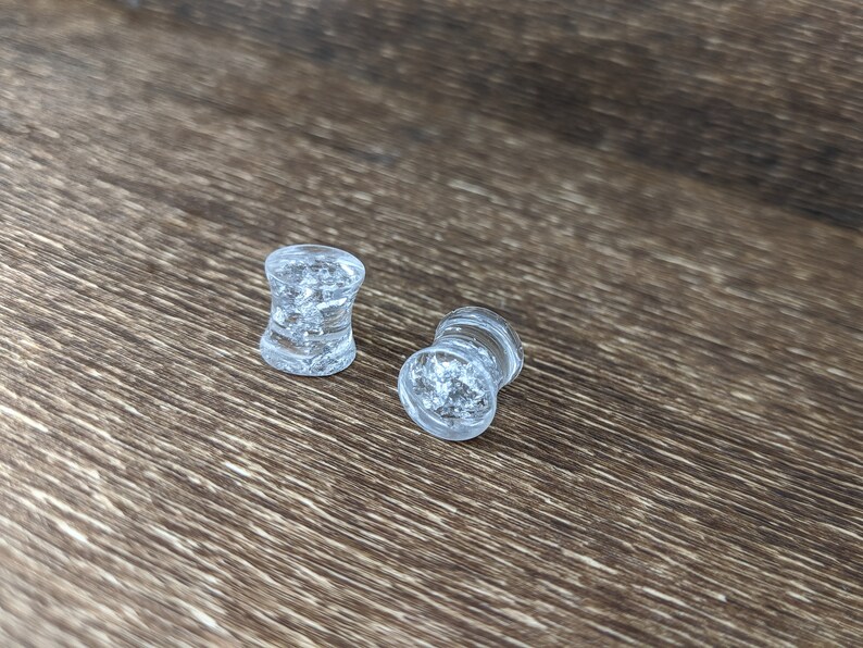 May include: Two clear glass ear gauges with a glittery, speckled texture. The gauges are on a brown wooden surface.