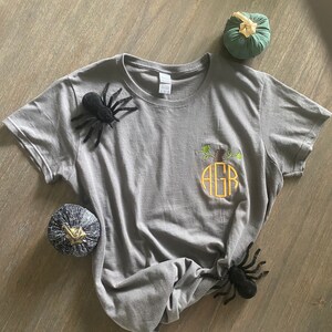 May include: A gray t-shirt with a pumpkin-shaped monogram embroidered on the front. The monogram is in orange and yellow and reads "AGR". There are three black spiders and two small pumpkins on the gray wood surface.