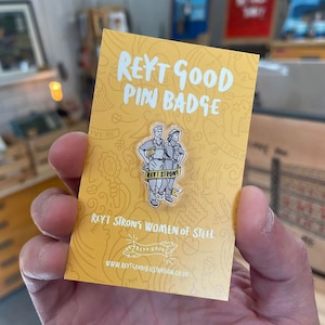 May include: A yellow card with the text "REYT GOOD PIN BADGE" and a cartoon image of two women wearing blue overalls and holding tools. The text "REYT STRONS WOMEN OF STEEL" is printed below the image. The text "REYT GOOD" is printed in a circle below the text.
