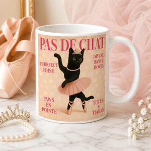 May include: White ceramic mug featuring a black cat in a pink tutu, tiara, and ballet shoes. The mug has the text "PAS DE CHAT" and other ballet-themed phrases. A pearl necklace and a tiara are also visible.