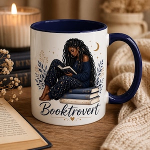 May include: White ceramic mug with a navy blue interior and handle. The mug features an illustration of a person reading, with the word "Booktrovert" below. The design includes stars, a crescent moon, and floral elements.