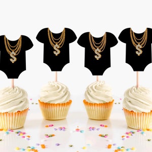 May include: Six black cupcake toppers shaped like baby onesies with gold chains and a diamond dollar sign pendant. The toppers are on white cupcakes with white frosting.