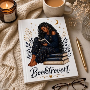 May include: A white journal with the word "Booktrovert" in script. The cover features an illustration of a person reading, surrounded by celestial and botanical elements. A candle, coffee cup, and pen are nearby.
