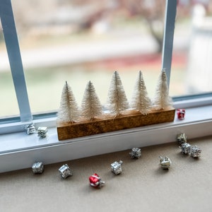 May include: A festive Christmas display. Five small, white bottle brush trees are set in a stained wooden base. Miniature silver and red gift boxes are scattered around the base, creating a festive scene.