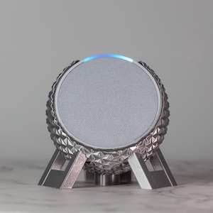 Puede incluir: Un altavoz esférico plateado con un patrón geométrico y un anillo de luz azul alrededor de la parte superior. El altavoz está en un soporte con tres patas.