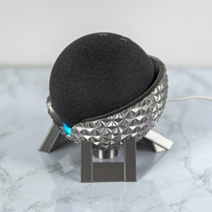 Puede incluir: Un altavoz inteligente esférico negro con un brillo azul claro, sentado en un soporte plateado con un patrón geométrico. El soporte tiene forma de esfera y tiene tres patas.