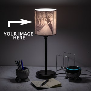 Puede incluir: Una lámpara de mesa negra con una pantalla cilíndrica blanca que presenta una foto en blanco y negro de una pareja. La lámpara está sobre una superficie gris con un cuaderno, un bolígrafo y un pequeño macetero gris con lápices.