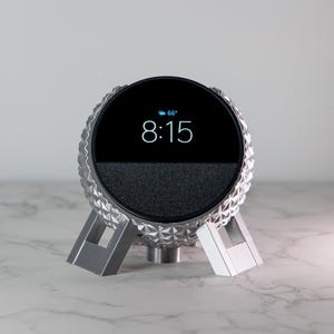 Puede incluir: Un altavoz inteligente plateado con un diseño esférico texturizado y una pantalla negra que muestra la hora 8:15 y la temperatura 68 grados. El altavoz está en un soporte plateado con tres patas.