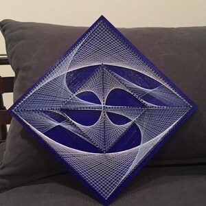 May include: A diamond-shaped piece of string art with white string woven into a geometric pattern on a dark blue background.