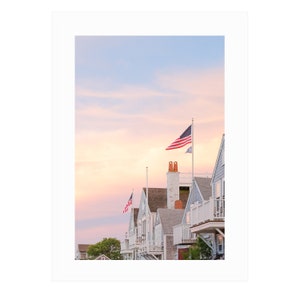 May include: A row of houses with light blue siding and white trim, each with a balcony. American flags fly from flagpoles on the rooftops against a pastel sky. The image evokes a sense of coastal charm and patriotism.