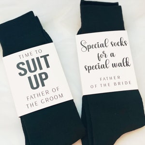 May include: Two pairs of black socks with white gift tags. The tag on the left sock reads "Time to Suit Up Father of the Groom". The tag on the right sock reads "Special socks for a special walk Father of the Bride".