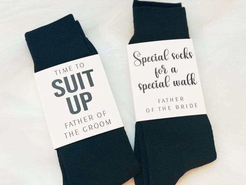 May include: Two pairs of black socks with white gift tags. The tag on the left sock reads "Time to Suit Up Father of the Groom". The tag on the right sock reads "Special socks for a special walk Father of the Bride".