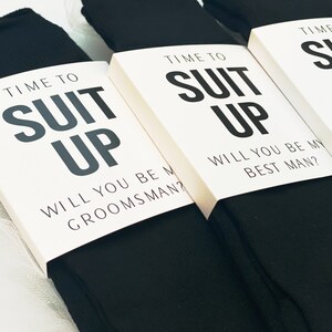 May include: Three pairs of black socks with white cards attached to them. The cards have the text "Time to Suit Up" and ask "Will you be my groomsman?" or "Will you be my best man?"