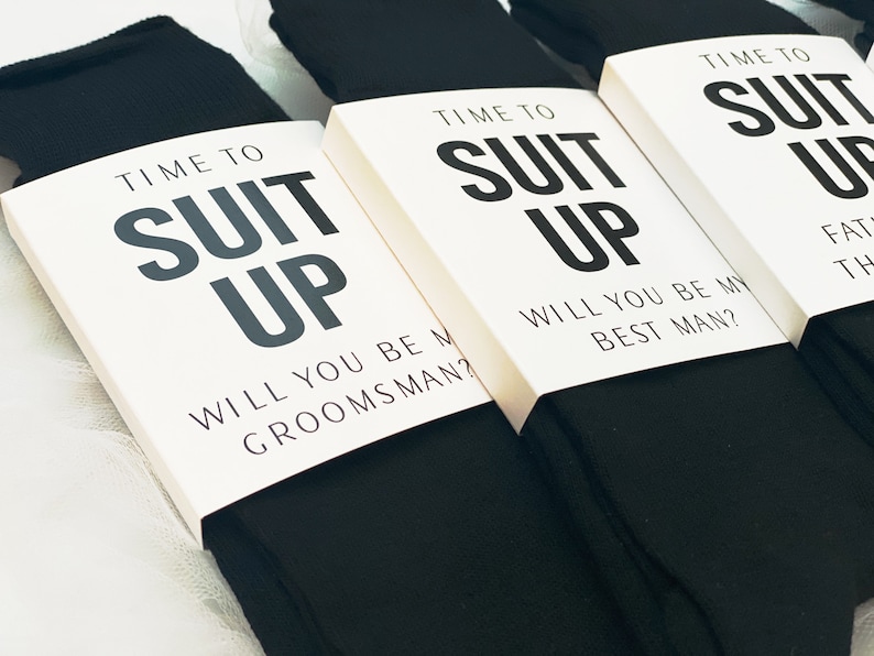May include: Three pairs of black socks with white cards attached to them. The cards have the text "Time to Suit Up" and ask "Will you be my groomsman?" or "Will you be my best man?"