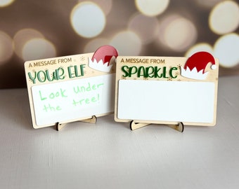 May include: Two wooden signs with a white dry erase surface and a festive elf hat design. The signs have a message that reads "A message from your elf" and "A message from sparkle". The sign on the left has the message "Look under the tree!" written on the dry erase surface.