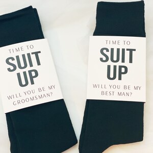 May include: Two pairs of black socks with white cards attached to them. The cards say "Time to Suit Up" and ask "Will you be my groomsman?" and "Will you be my best man?"