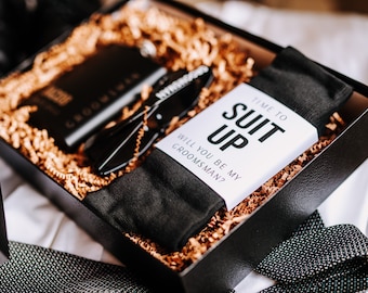 May include: A black gift box filled with brown crinkle paper. The box contains a black pair of socks, a black flask, a pair of black sunglasses, and a white card with the text "Time to Suit Up. Will you be my groomsman?"