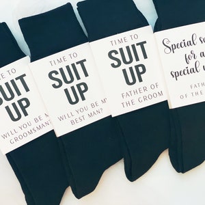 May include: Four pairs of black socks with white tags attached. The tags have the text "Time to Suit Up" with different messages below. One tag says "Will you be a groomsman?", another says "Will you be my best man?", another says "Father of the Groom", and the last one says "Special socks for a special walk Father of the Bride."