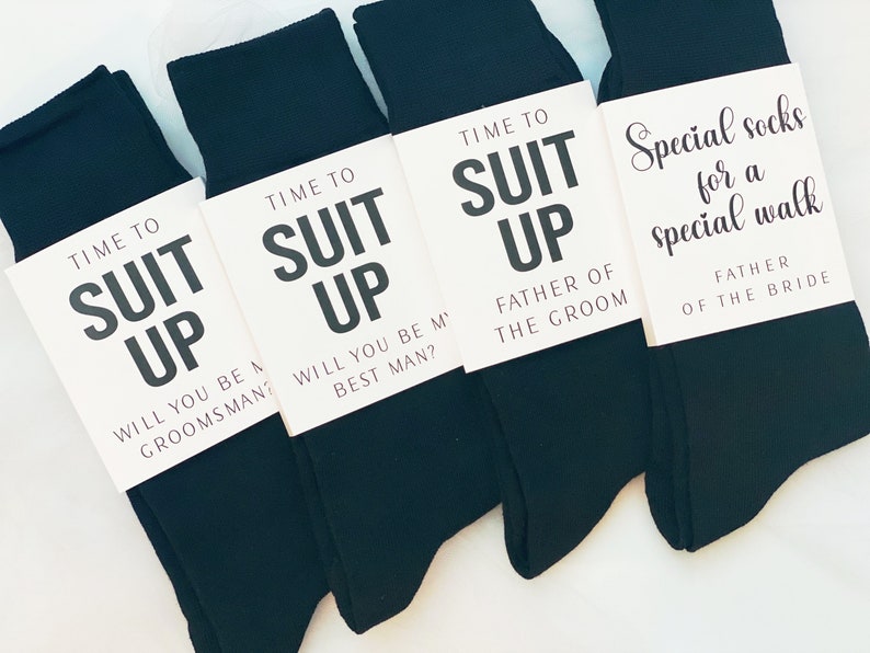 May include: Four pairs of black socks with white tags attached. The tags have the text "Time to Suit Up" with different messages below. One tag says "Will you be a groomsman?", another says "Will you be my best man?", another says "Father of the Groom", and the last one says "Special socks for a special walk Father of the Bride."