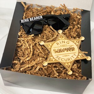 May include: A black gift box filled with brown crinkle paper. The box contains a pair of black sunglasses with the text "RING BEARER" on the frame and a wooden sheriff's badge with the text "RING SAWYER SECURITY".