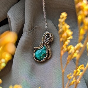 May include: A handcrafted pendant necklace featuring a vibrant green gemstone. The stone is encased in a silver and gold-toned wire wrap, suspended from a delicate silver chain. The background is a soft, muted green fabric.