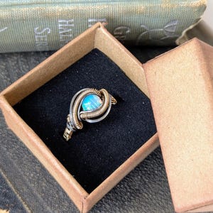 May include: A ring with a blue opal stone set in a gold and silver wire wrapped design. The ring is in a small brown box.