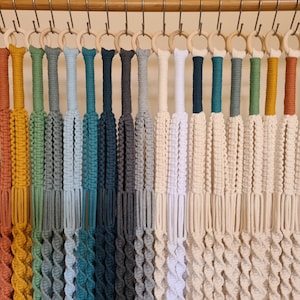 May include: A collection of 20 macrame plant hangers in various colours, including red, orange, yellow, green, blue, and white. Each hanger features a wooden ring at the top and a spiral design.