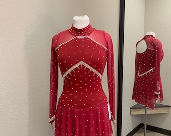 May include: A red figure skating dress with long sheer sleeves and a high neckline. The dress is adorned with white rhinestones and a flared skirt. A matching dress is reflected in a nearby mirror.