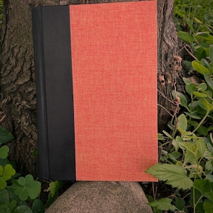 May include: A hardback book with a black spine and a textured, coral-coloured cover. The book is resting on a rock, with a tree trunk and greenery in the background. The book's cover has a woven texture.
