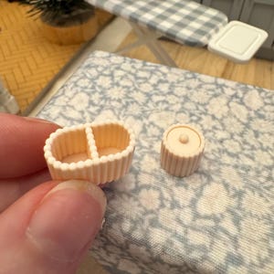 May include: Miniature peach-coloured dollhouse accessories. The set includes a divided basket and a small, round container. The basket has a ribbed design and a beaded edge. The container has a small knob on top. These items are likely for a dollhouse kitchen.