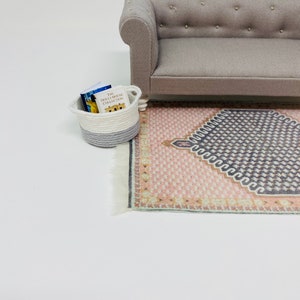 Pu&ograve; includere: Scena di casa delle bambole in miniatura con un divano grigio, un tappeto fantasia nei toni del rosa e del grigio e un piccolo cestino con libri. Il cestino &egrave; bianco e grigio. I libri sono etichettati "The Dollhouse Collection."
