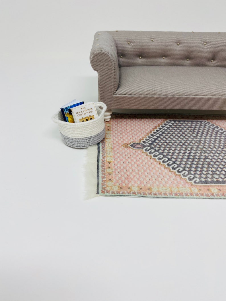 Pu&ograve; includere: Scena di casa delle bambole in miniatura con un divano grigio, un tappeto fantasia nei toni del rosa e del grigio e un piccolo cestino con libri. Il cestino &egrave; bianco e grigio. I libri sono etichettati "The Dollhouse Collection."