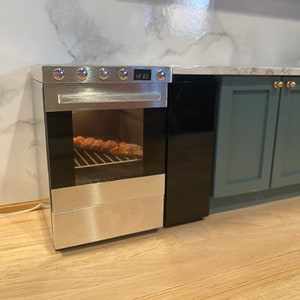 May include: A miniature kitchen stove with a brushed metal finish, a black glass oven door, and a digital display showing 12:30. The oven interior contains a small rack with baked goods. It is positioned next to dark teal cabinets with gold knobs.