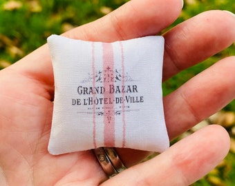May include: A small white pillow with pink stripes and the text "Grand Bazar de l'Hotel de Ville" printed on it.