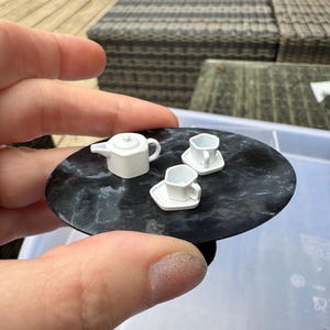 Puede incluir: Juego de té en miniatura de porcelana blanca, que incluye una tetera y dos tazas con platillos, dispuesto sobre una pequeña bandeja redonda negra con efecto mármol. El juego se sostiene en una mano, lo que sugiere una pequeña escala.