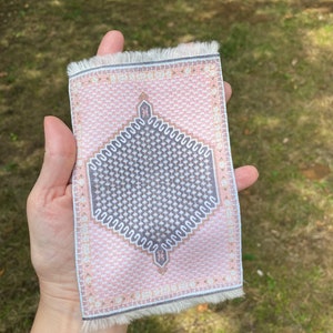 Pu&ograve; includere: Un piccolo tappeto rettangolare con un motivo geometrico rosa e grigio. Il tappeto presenta un disegno centrale a forma di diamante e bordi sfrangiati. &Egrave; tenuto in mano su uno sfondo verde sfocato.