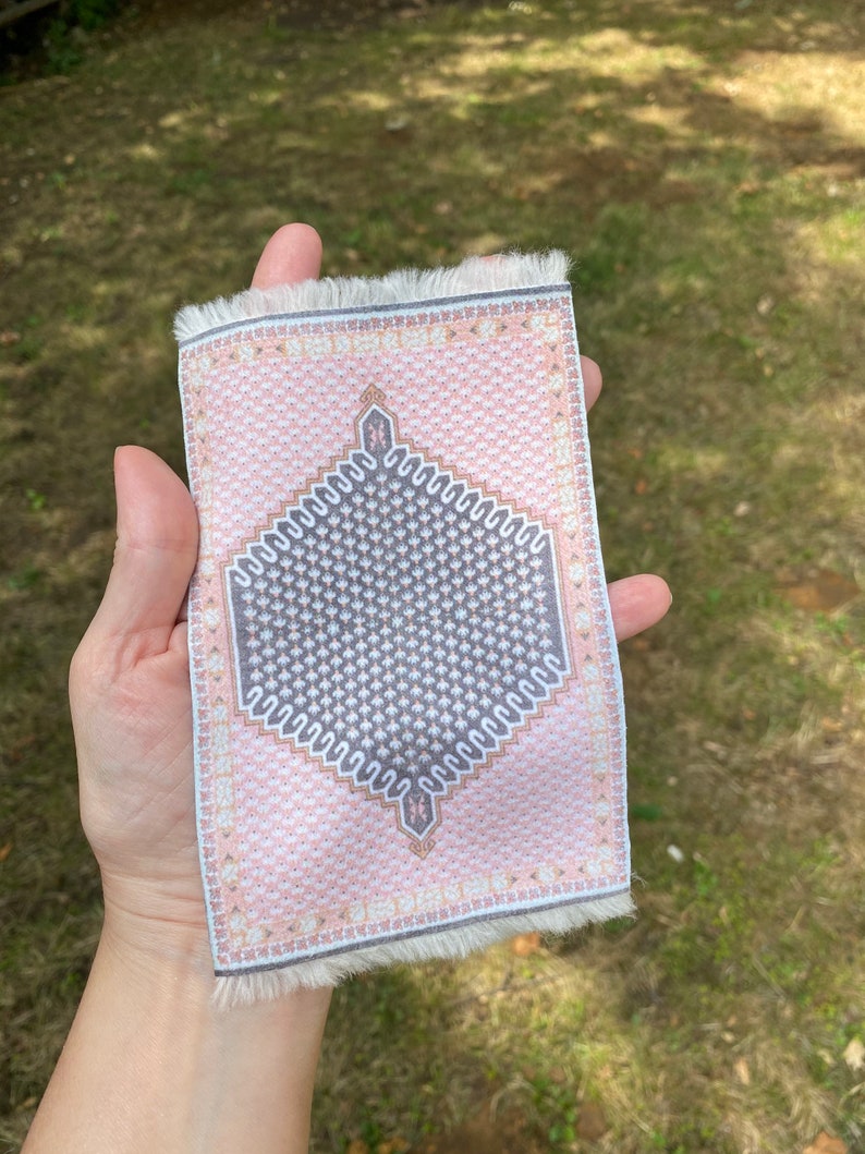 Pu&ograve; includere: Un piccolo tappeto rettangolare con un motivo geometrico rosa e grigio. Il tappeto presenta un disegno centrale a forma di diamante e bordi sfrangiati. &Egrave; tenuto in mano su uno sfondo verde sfocato.