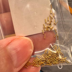 Könnte beinhalten: Eine durchsichtige Plastiktüte gefüllt mit einer großen Menge kleiner, goldfarbener Metall-Stifte.