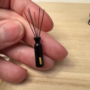 May include: A small, black vase with thin, dark rods extending upwards. A small, vertical yellow stripe is on the side of the vase. The object is held in a person's hand against a light wood surface.